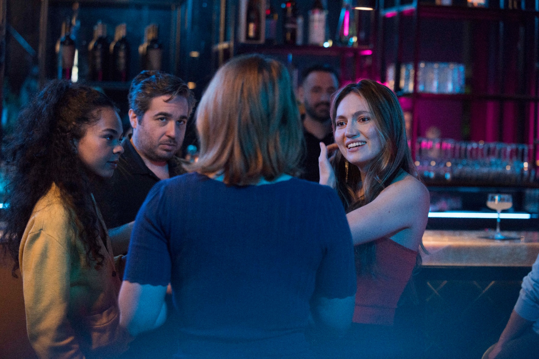 A group gathers around the bar in moody neon light.