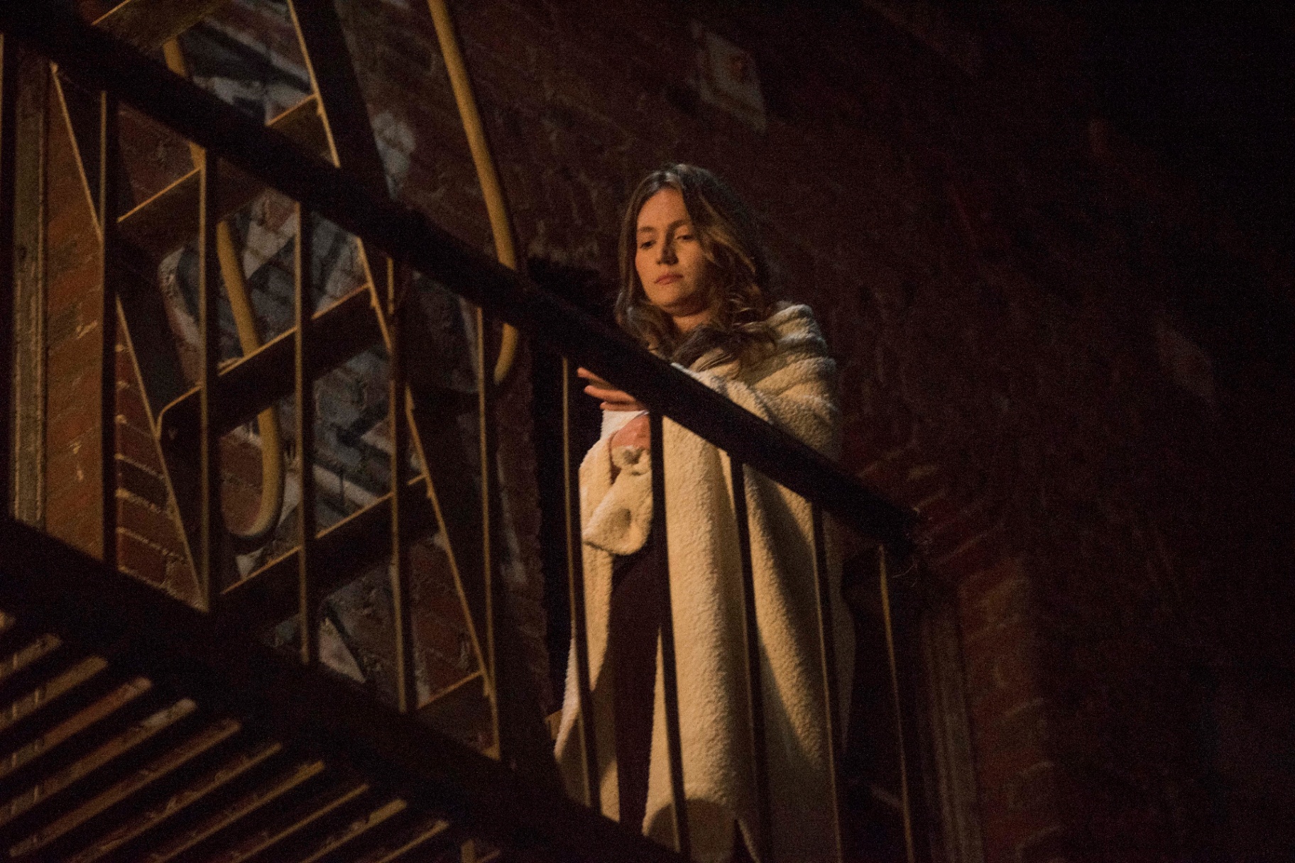 Amanda on a fire escape at night, wrapped in a cream coat.