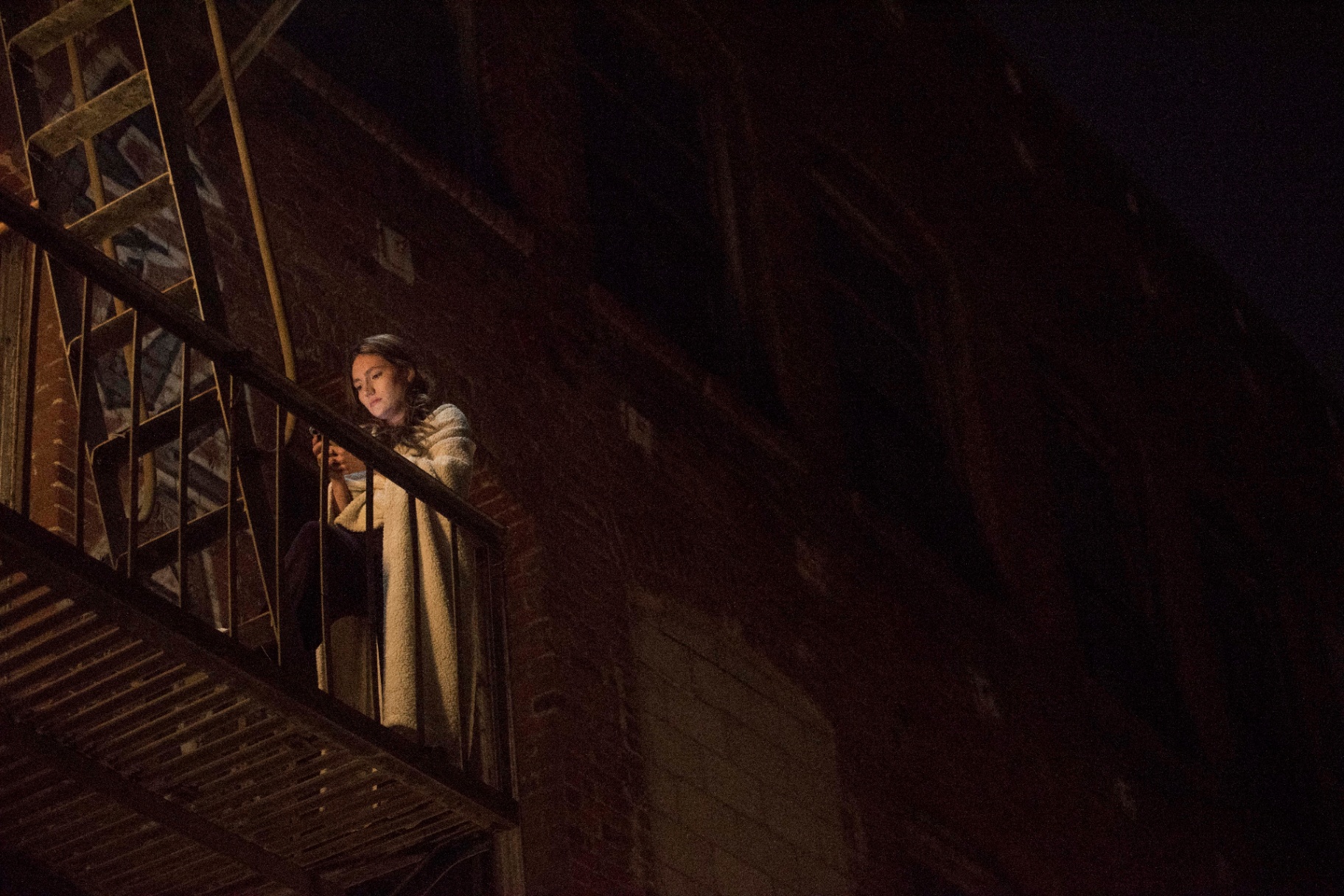 A woman sits on a fire escape at night, lit by her phone.
